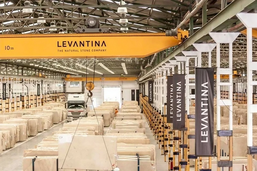 Almacén industrial de Levantina con grúa elevadora de 10 toneladas manipulando bloques de granito y cuarcita Naturamia, filas de tablas de piedra natural empaquetadas y banners de la marca, destacando la cadena de suministro premium para encimeras en Málaga