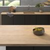 Encimera Arklam Arcane Camel madera con bol de peras, fregadero y gabinetes negros en cocina moderna.