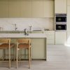 Cocina moderna con encimera Dekton Keena blanca, gabinetes verdes suaves, taburetes madera y electrodomésticos integrados.