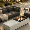 Terraza lounge con brasero Dekton Nara, sillones grises, manta y vegetación en entorno soleado.