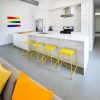 Isla blanca Neolith Arctic White con taburetes amarillos, arte arcoíris y electrodomésticos en cocina abierta.