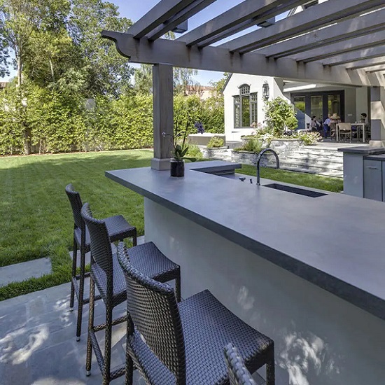 Barra exterior Neolith Aspen Grey gris con taburetes, fregadero y jardín verde al fondo.