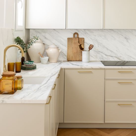 Cocina con encimera Neolith Azure blanca veteada, gabinetes beige, grifo dorado y decoraciones naturales.