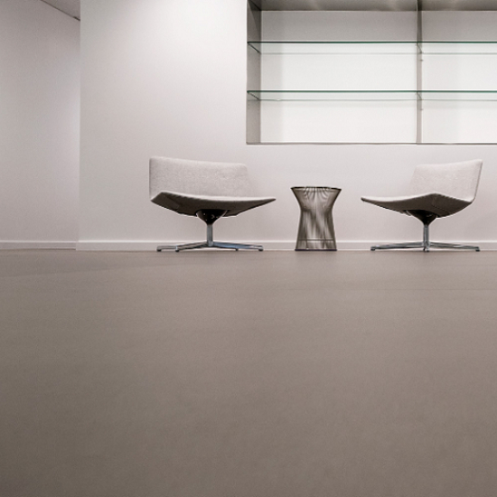 Sala minimalista con sillas blancas y mesa plateada sobre suelo Neolith Barro terracota.