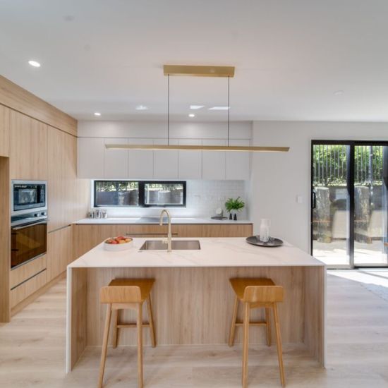 Cocina con isla Blaker Statuario Gold blanca veteada, taburetes de madera, luz lineal y gabinetes blancos en ambiente espacioso.