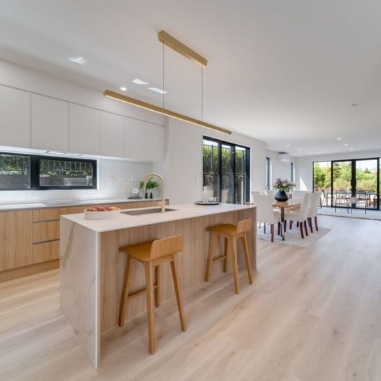 Cocina elegante con isla Blaker Statuario Gold blanca, sillas teal, gabinetes oscuros y piso marrón en diseño lujoso.